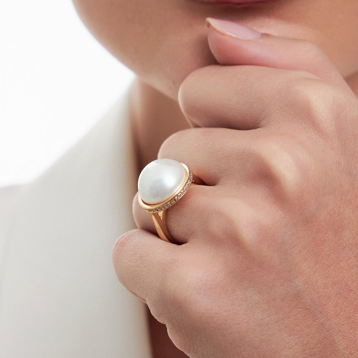 A close-up of a model wearing a gold ring with a large white pearl and small clear stones in a halo setting.