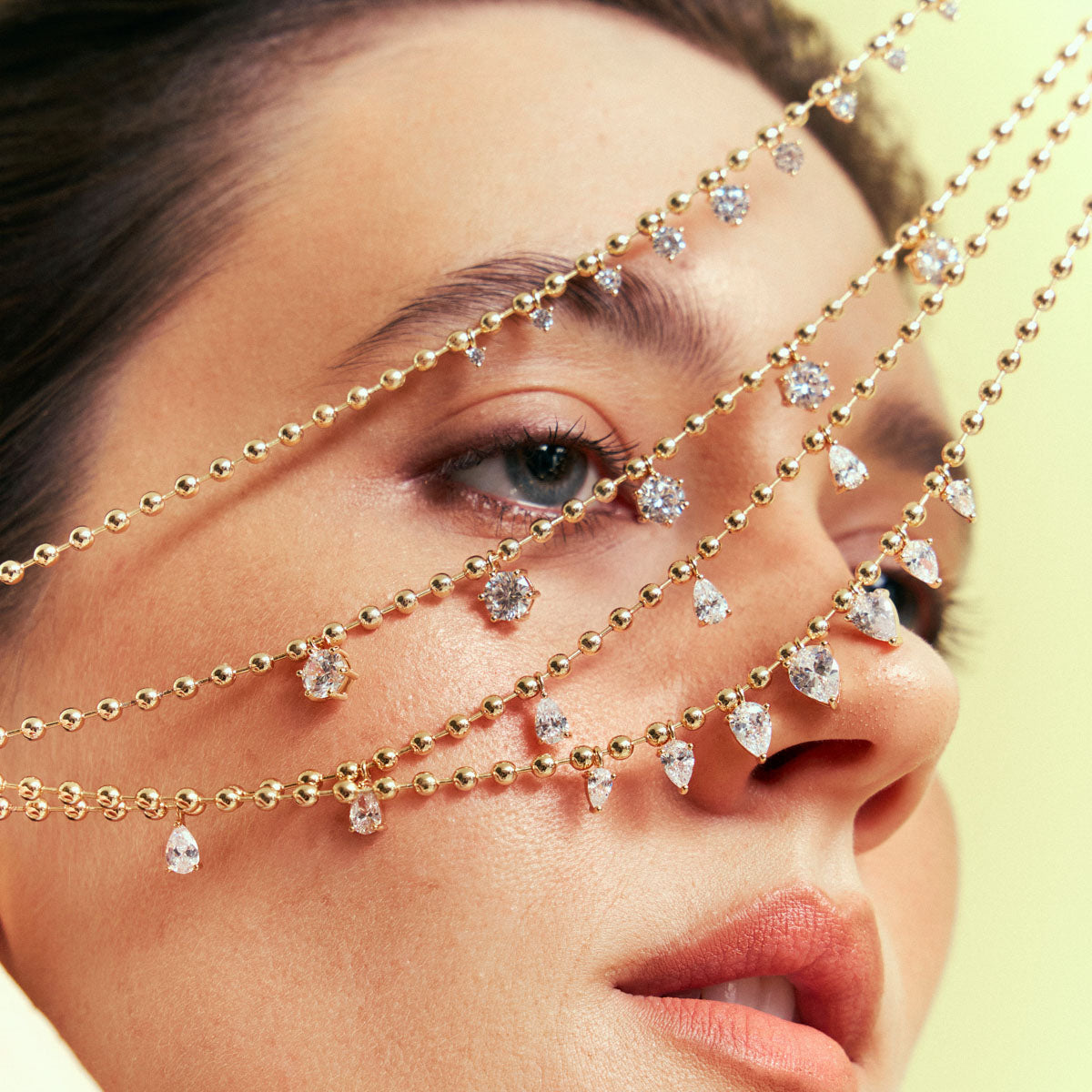 A close-up of a model's face wearing delicate gold chain headpieces adorned with clear teardrop and round gemstones across the forehead, eyes, and nose.