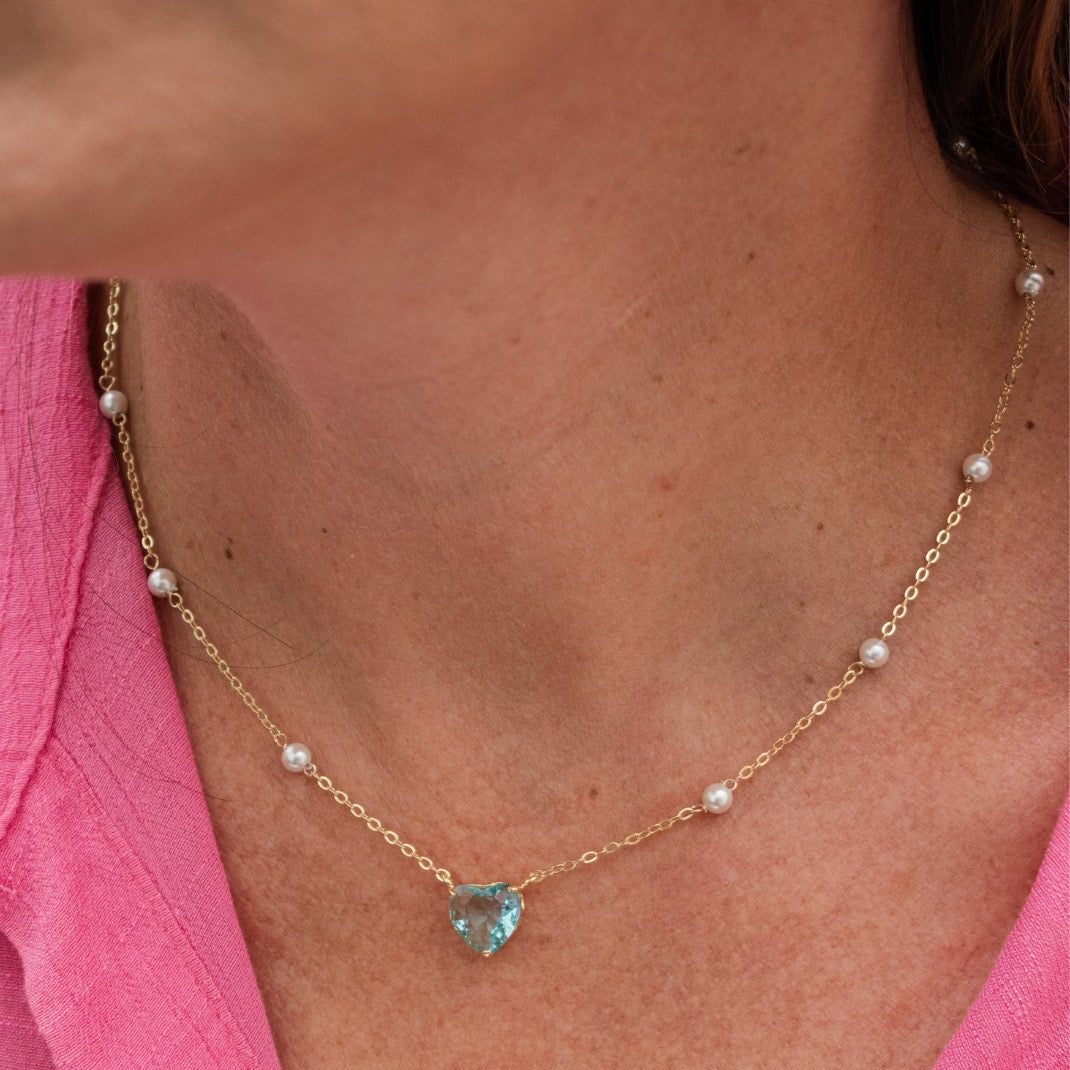 A close-up of a woman wearing a delicate gold chain necklace with small white pearls spaced along the chain and a heart-shaped light blue gemstone pendant at the center. She is wearing a bright pink top.