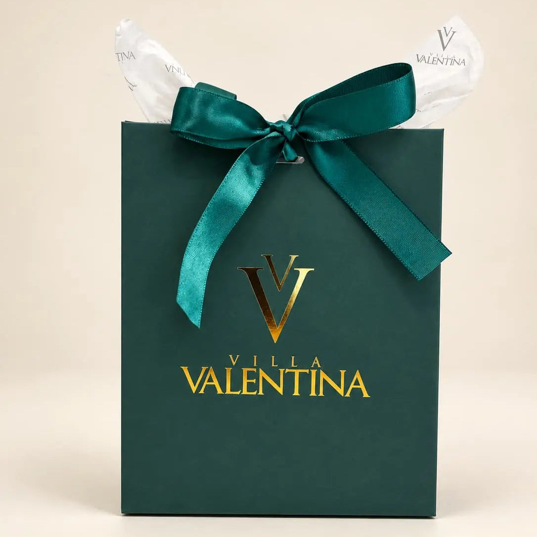 A dark green Villa Valentina gift bag with a metallic gold logo and the brand name printed on the front, tied with a large dark green satin ribbon and white tissue paper, set against a plain beige background.