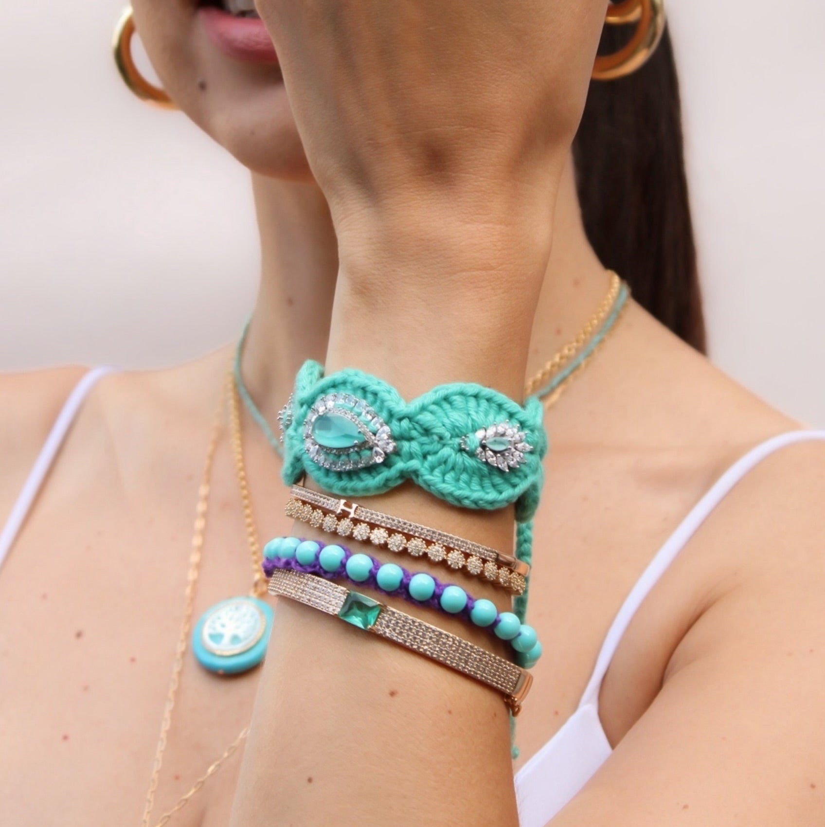 A close-up lifestyle shot of a woman wearing a stack of turquoise and blue beaded bracelets, a turquoise beaded necklace, and a handmade crocheted bracelet with rhinestones on her wrist.