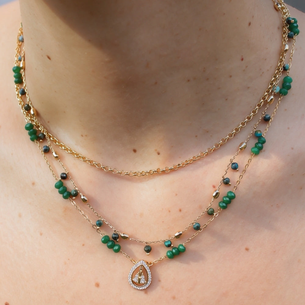 A close-up of a model's neck and collarbone wearing a layered gold necklace with green bead accents and a teardrop-shaped pendant.