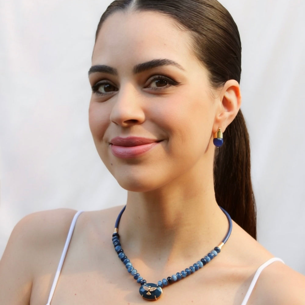 A woman with dark hair in a ponytail wearing a blue beaded necklace and matching drop earrings, posing against a light off-white background.