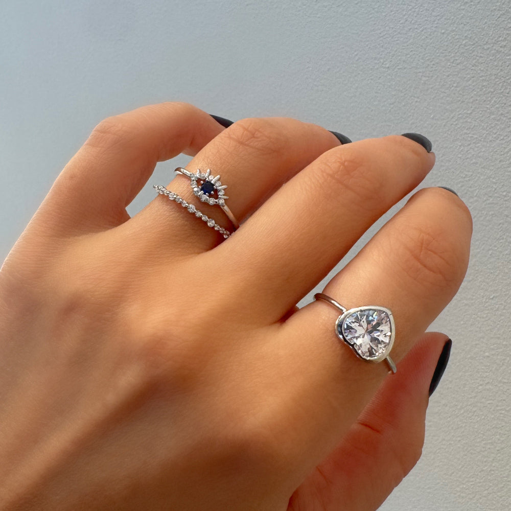 A close-up of a hand wearing three silver rings: an eye-of-providence ring with a dark blue oval stone, a delicate pavé band ring, and a silver ring featuring a heart-shaped clear gemstone.