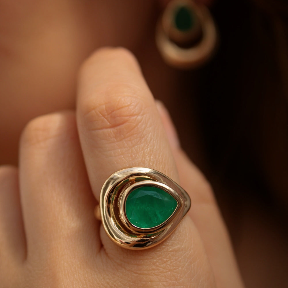 A gold ring with a teardrop-shaped green emerald stone, worn on a person's finger. A matching green and gold drop earring is visible in the blurred background.