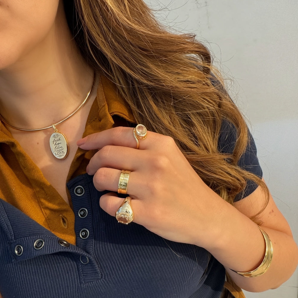 A close-up lifestyle shot of a woman wearing multiple gold-toned rings and a gold pendant necklace over a navy blue ribbed polo shirt and a mustard yellow collared undershirt.
