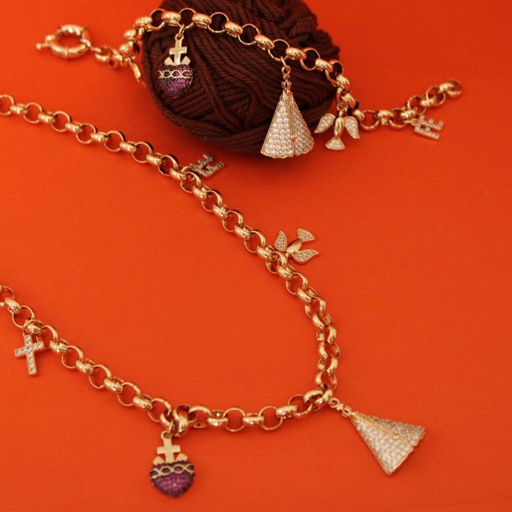 A gold-tone chain necklace with multiple charms, including a heart pendant with purple enamel, a small cross, and textured cone pendants, displayed on a bright orange background next to a ball of dark brown yarn.