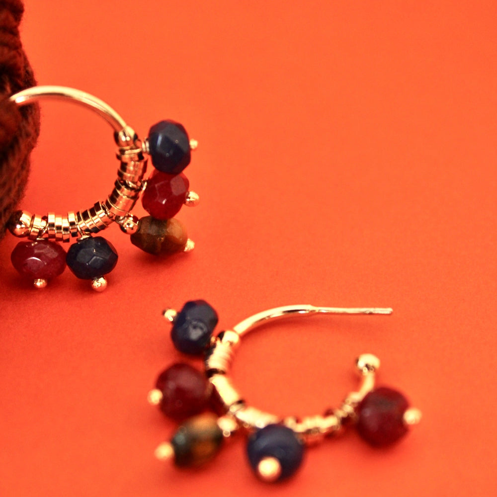 Gold hoop earrings with dangling faceted natural jade in dark red, navy blue, and brown, displayed on a vibrant orange background.