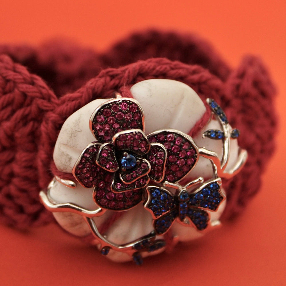 A close-up of a chunky knit cuff bracelet in deep burgundy red, featuring a large central floral and bow motif embellished with silver-toned metal, white stones, and dark red and blue rhinestones, set against a solid orange background.