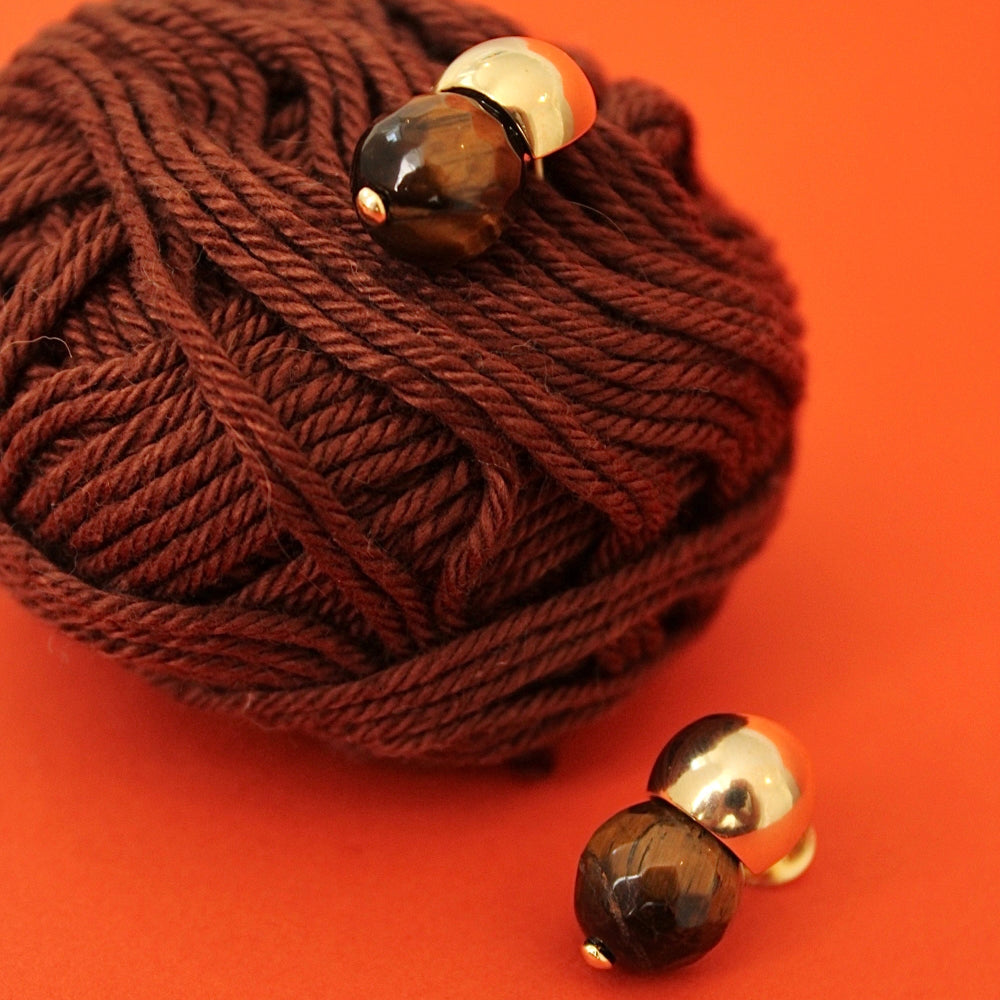 A pair of gold-toned double-bead earrings with faceted brown beads, displayed on a ball of dark brown yarn against a solid orange background.