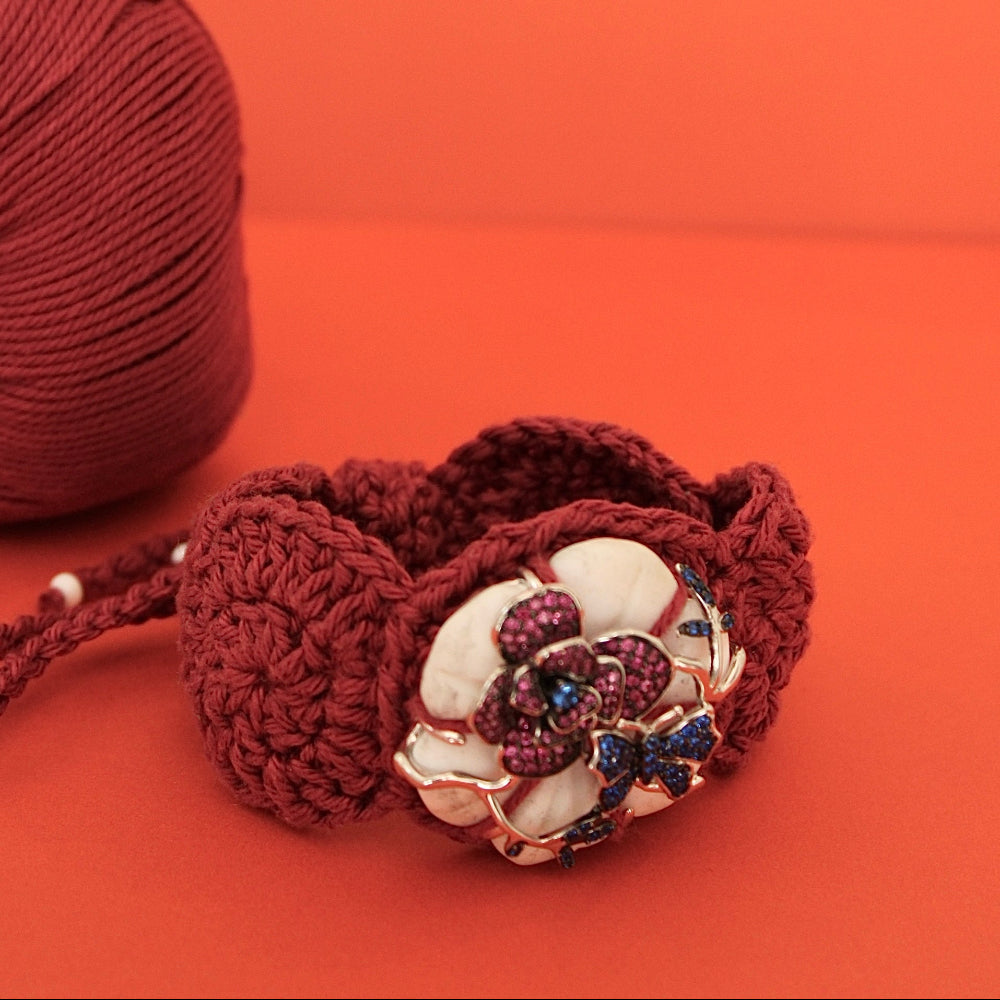 A close-up of a dark red crocheted bracelet with a large decorative flower charm featuring white petals and red jewel-like accents, set against a solid orange background with a matching red yarn ball partially visible on the left.