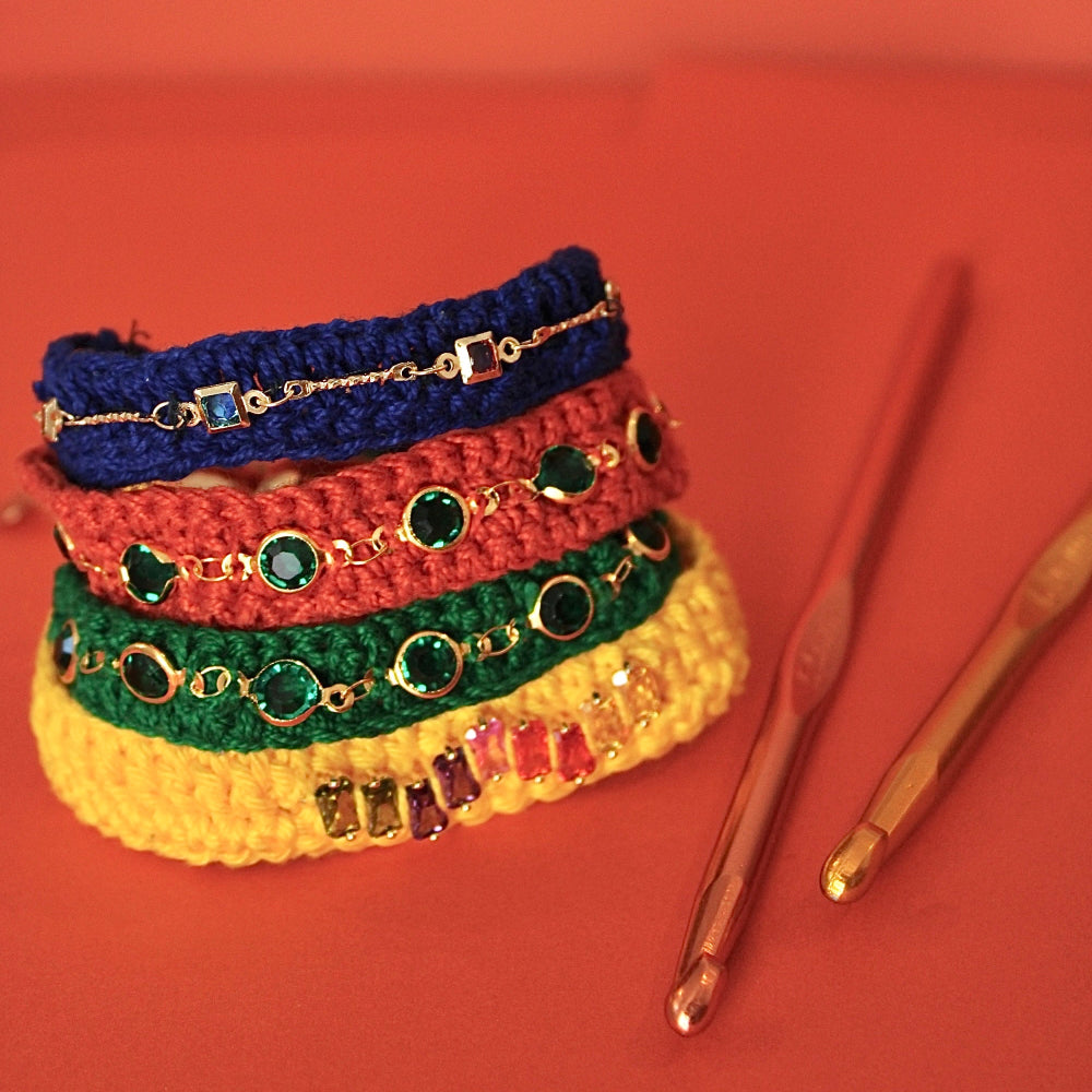 A stack of four colorful crocheted headbands with gold chain and rhinestone embellishments, displayed on a warm orange-red background next to two crochet hooks.