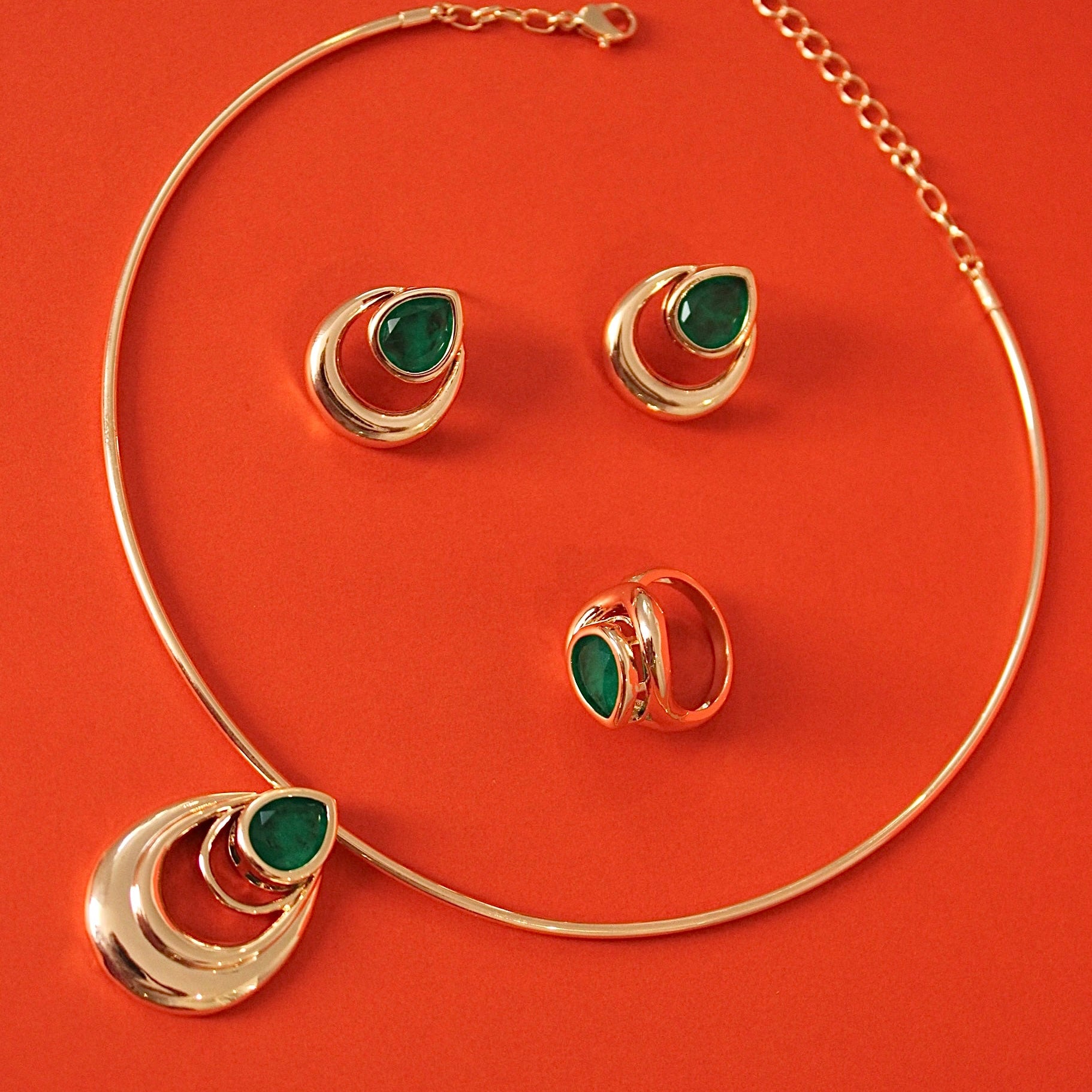 Gold jewelry set featuring a choker necklace, matching earrings, and a ring, each adorned with a teardrop-shaped green stone, displayed on a vibrant orange-red background.