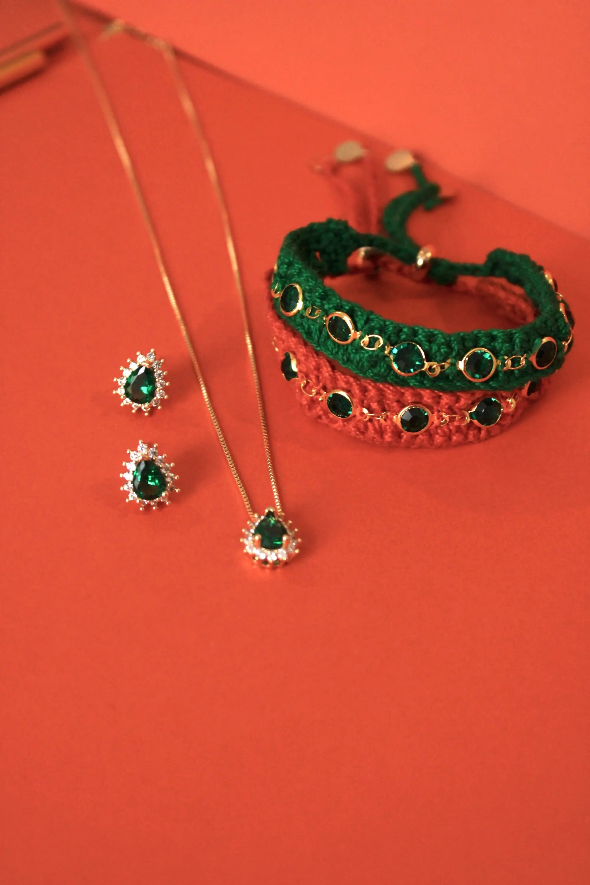 A jewelry set featuring a gold necklace with a green teardrop pendant, matching green teardrop stud earrings, and a green braided bracelet with red woven accents and gold circular details, arranged on a solid red background.