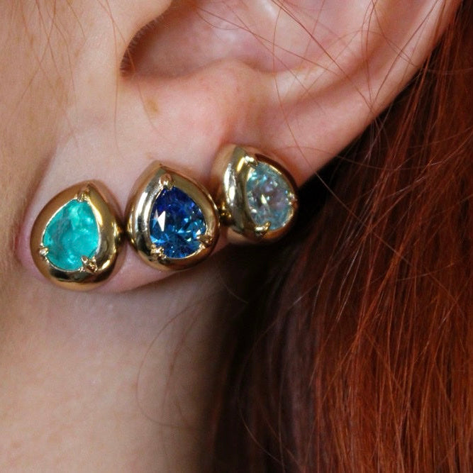 A close-up of a person's ear wearing three gold teardrop-shaped stud earrings with turquoise, blue, and clear gemstones.