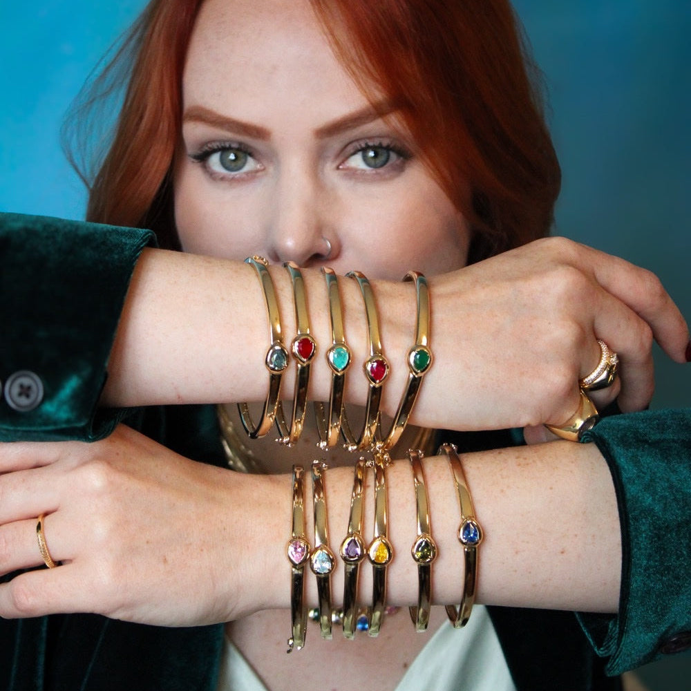 A close-up lifestyle shot of a woman with red hair wearing multiple gold bangle bracelets stacked on her arms. Each bracelet features a different colored gemstone (red, green, blue, yellow, pink, and purple) set in a polished gold band.
