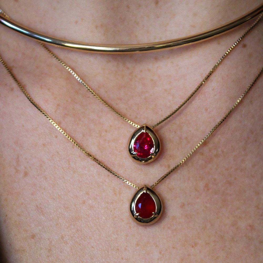 A close-up of a person's neck and chest wearing a layered necklace set. The set includes a wide gold choker, two delicate gold chains with teardrop-shaped red gemstone pendants, and a thin gold chain with a small gold oval pendant.