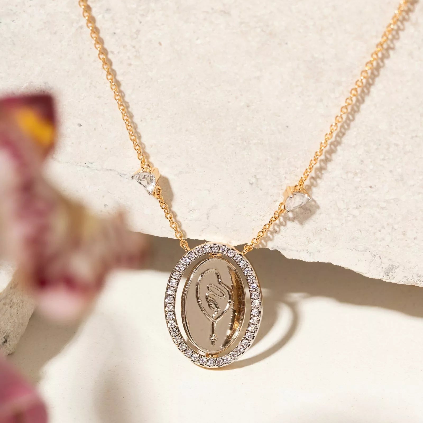 A gold oval pendant necklace with a clear crystal halo and a central gold medallion, displayed on a textured white stone surface with soft pink flowers in the foreground.