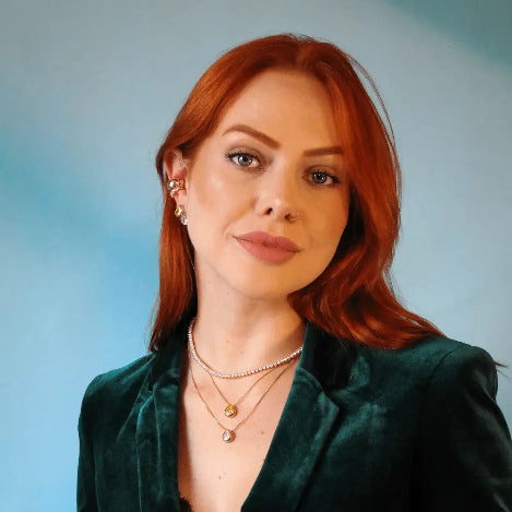 A close-up portrait of a woman with red hair wearing a dark green velvet blazer and layered gold necklaces, set against a soft light blue background.