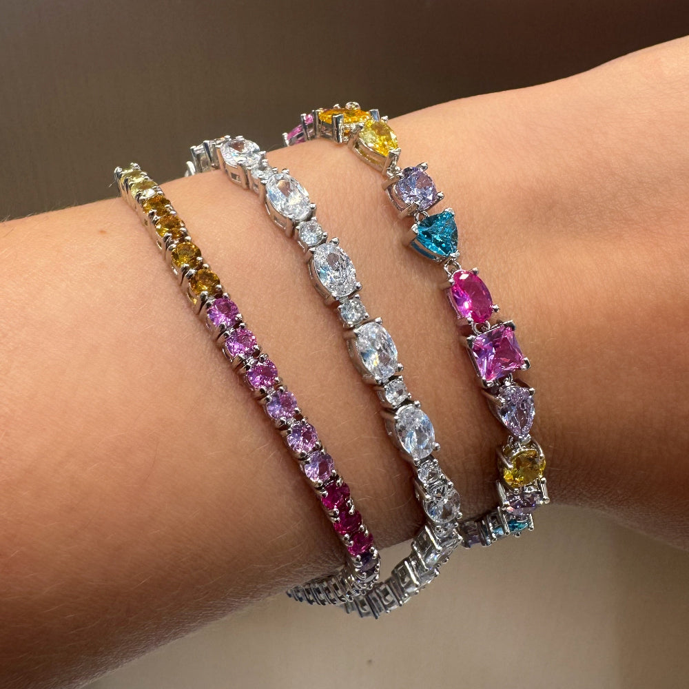 Three silver tennis bracelets with colorful gemstones are shown worn on a person's wrist. The bracelets feature yellow, pink, and blue stones.