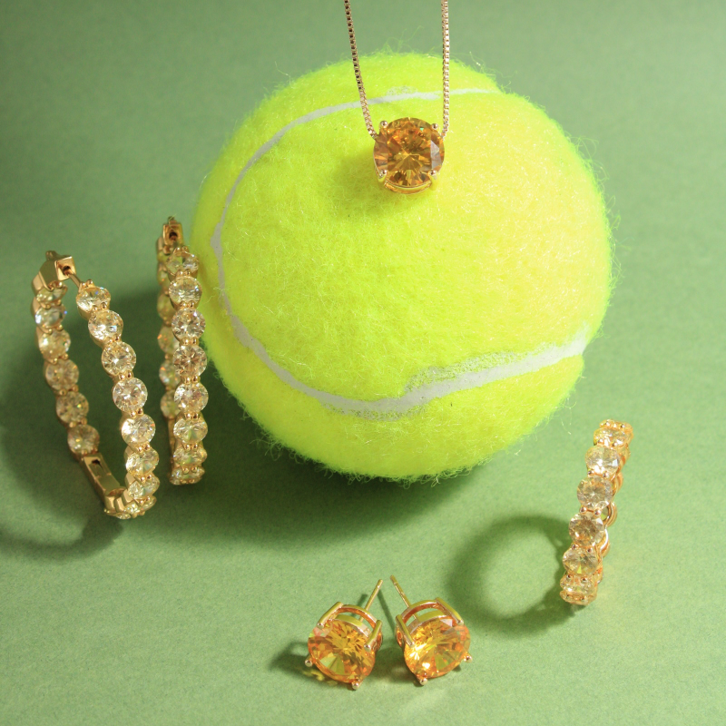 A tennis ball necklace with a round amber gemstone pendant, displayed on a green background alongside matching gold-tone jewelry including hoop earrings, stud earrings, and a tennis ball pendant.