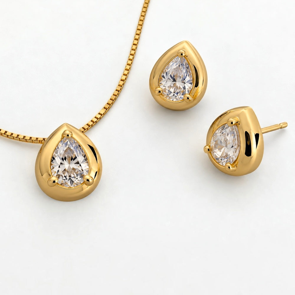Gold jewelry set featuring a teardrop-shaped pendant necklace, matching stud earrings, and a post earring, each set with a clear teardrop-cut gemstone on a white background.