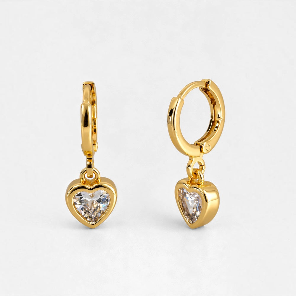 A pair of gold hoop earrings with dangling heart-shaped clear crystal charms, shown on a light gray background.