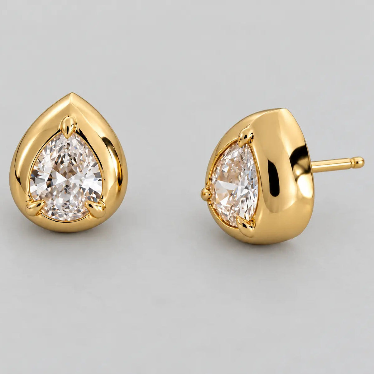 A pair of gold teardrop stud earrings with pear-cut clear gemstones, shown on a neutral light gray background. One earring is facing forward to show the setting and gemstone, while the other is shown in profile with its post visible.