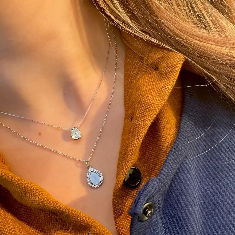 A close-up lifestyle shot of a person wearing two delicate silver necklaces: a shorter chain with a teardrop-shaped clear stone and a longer chain with a teardrop-shaped light blue stone surrounded by small clear stones. The person is wearing an orange shirt layered under a blue corduroy button-up shirt.