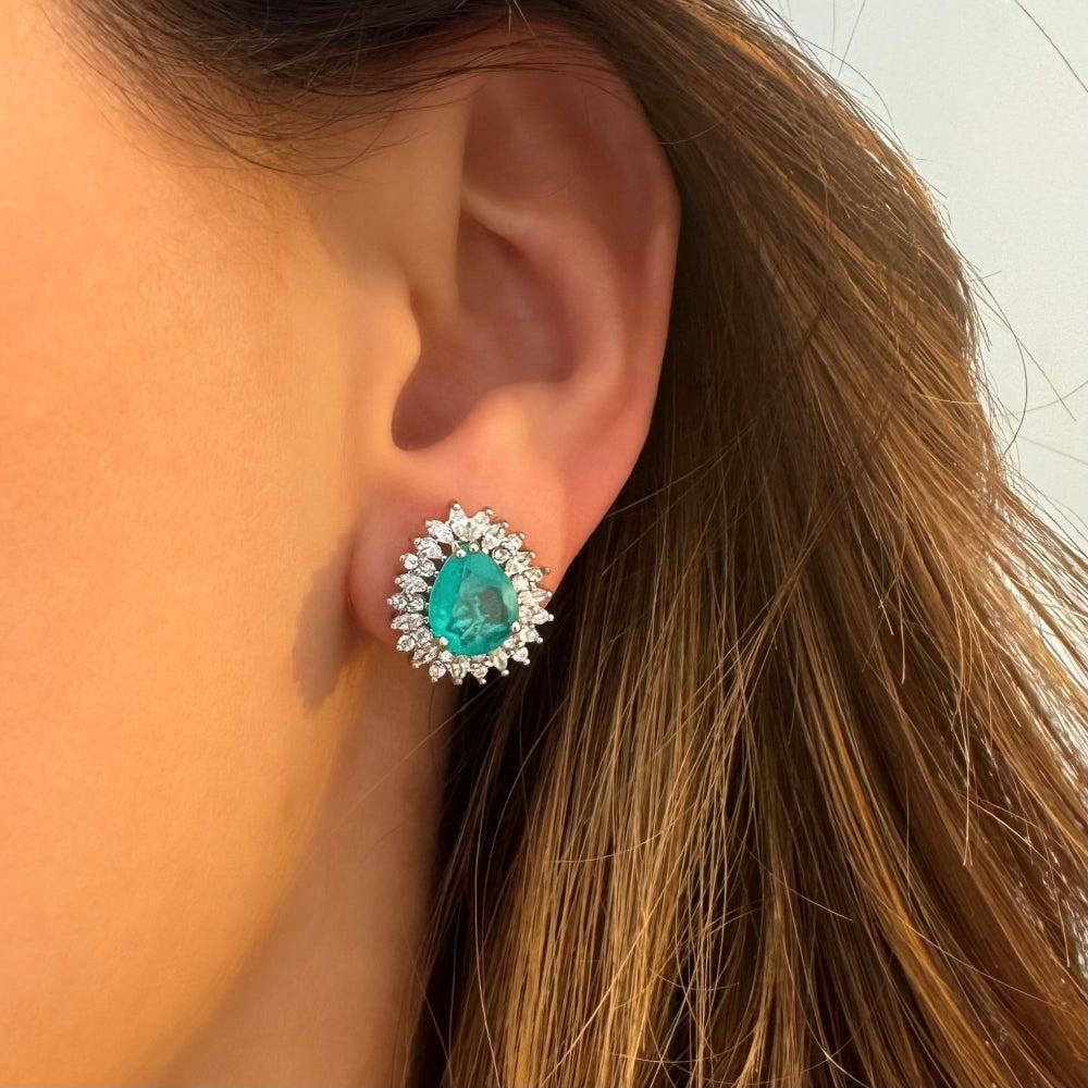 A close-up of a person's ear wearing a stud earring with a central turquoise-blue oval gemstone surrounded by a halo of small clear stones.