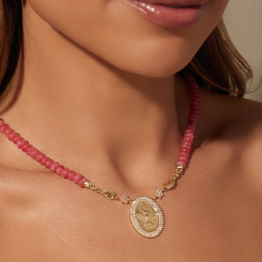 A close-up of a woman wearing a pink beaded necklace with a gold oval pendant and a matching pink beaded earring.