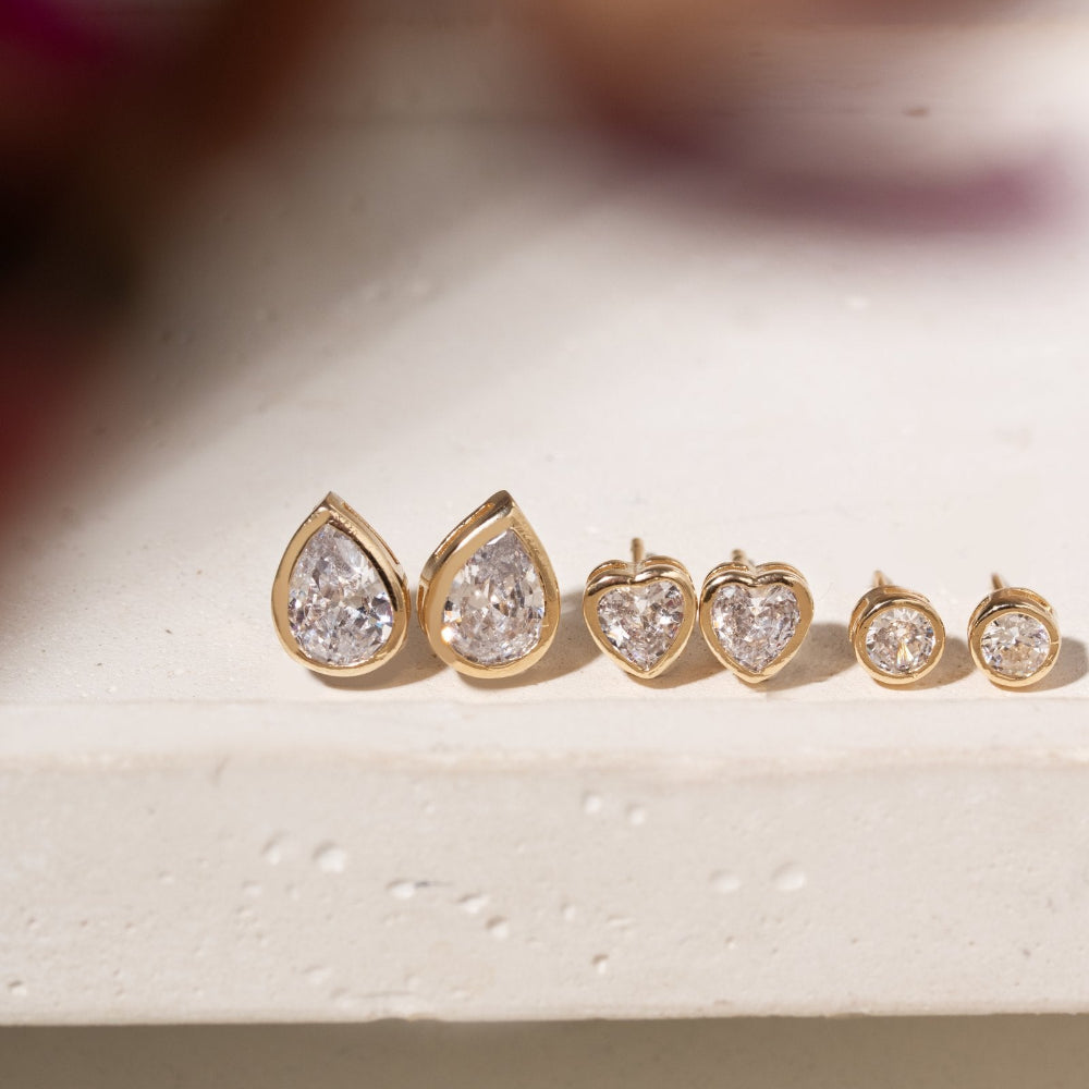 A set of five gold stud earrings with clear gemstones, arranged in descending sizes. The earrings feature pear-shaped stones on the largest pair and heart-shaped stones on the smaller pairs, displayed on a textured white surface.