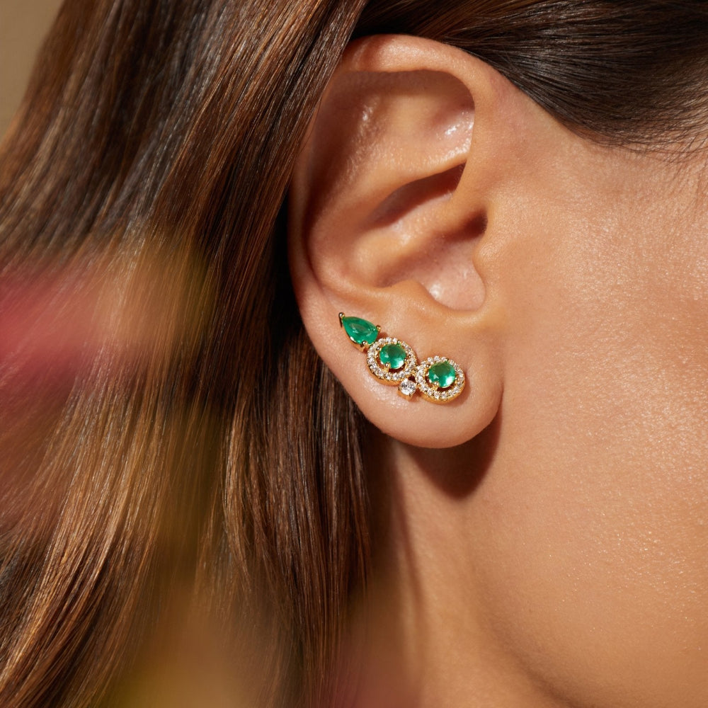 A close-up of a woman's ear wearing a pair of gold ear cuffs decorated with green emerald-cut gemstones and small clear crystals.