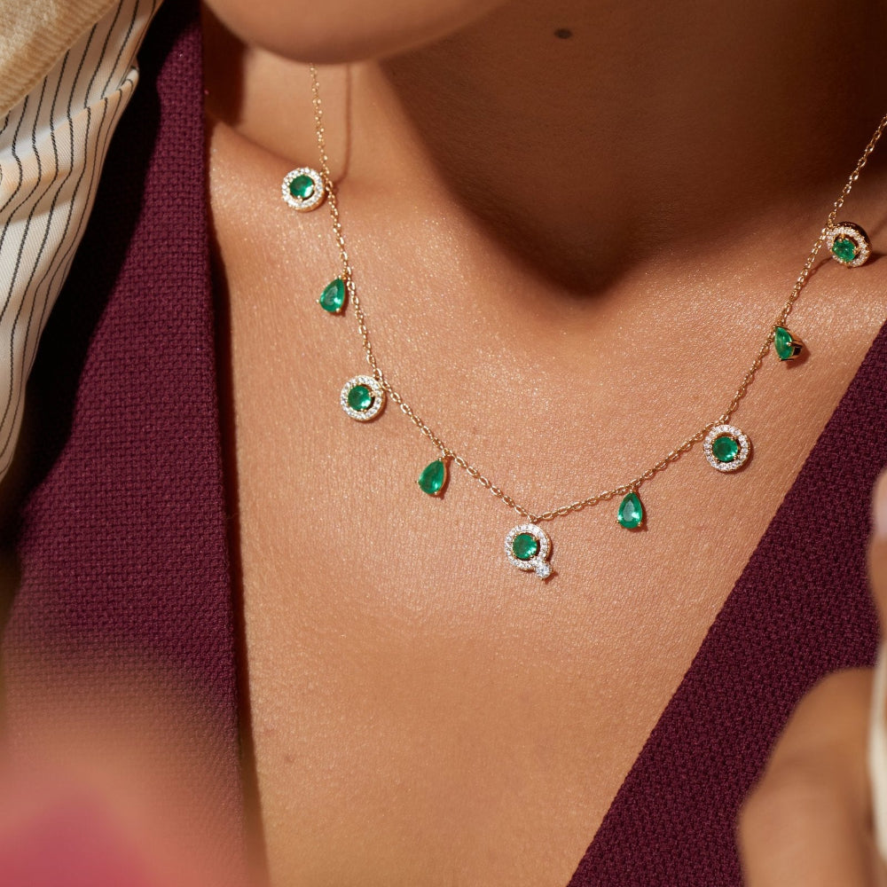 A close-up lifestyle shot of a model wearing a delicate gold necklace adorned with green emerald-cut gemstones and small clear stones.