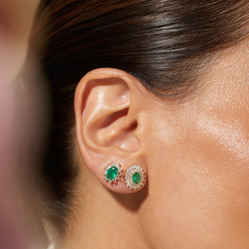 Close-up of a model's ear wearing a pair of oval stud earrings with green emerald-cut centers surrounded by small clear stones in a gold-tone setting.