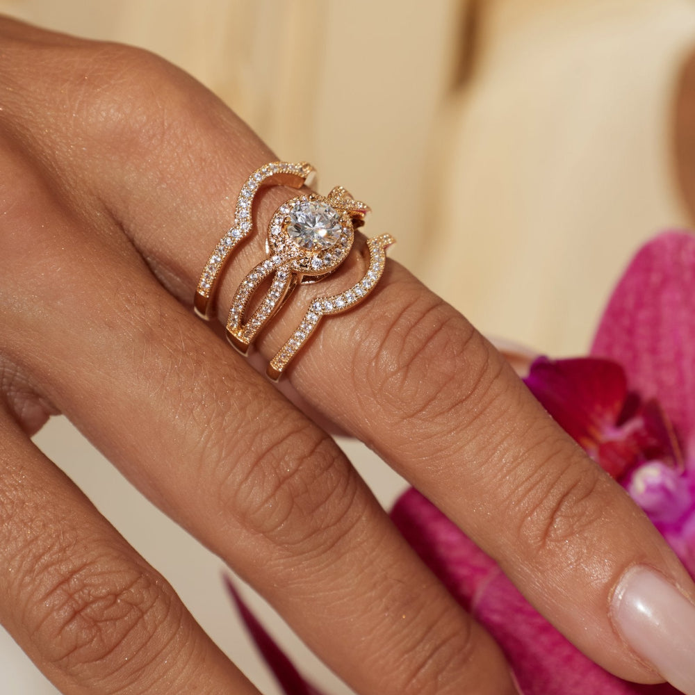 A close-up of a hand wearing a rose gold ring set with a round-cut center stone and curved pavé bands, styled with a pink orchid flower against a soft cream background.