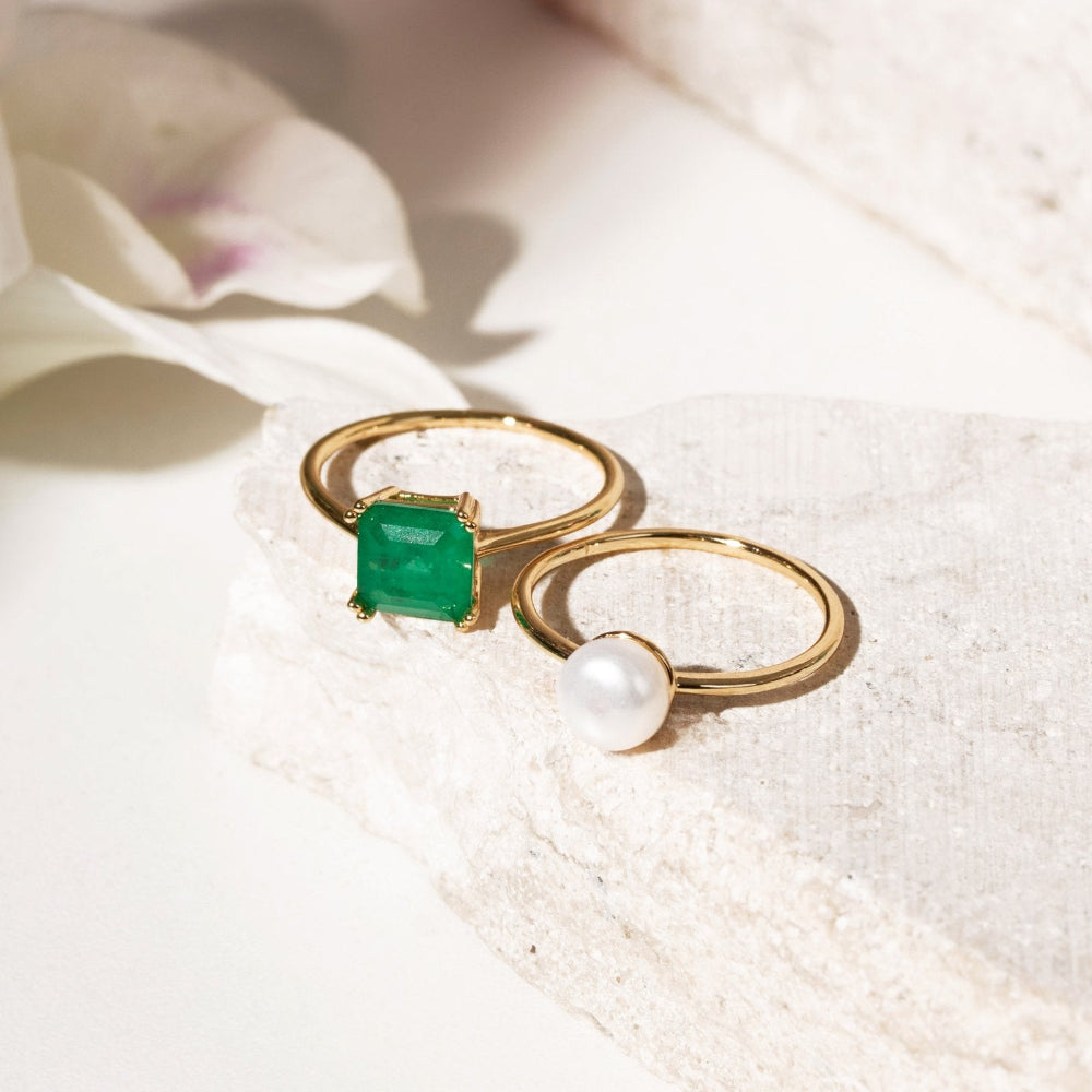 Two gold rings displayed on a textured white stone surface. One ring features a square-cut emerald green gemstone, and the other features a round white pearl.