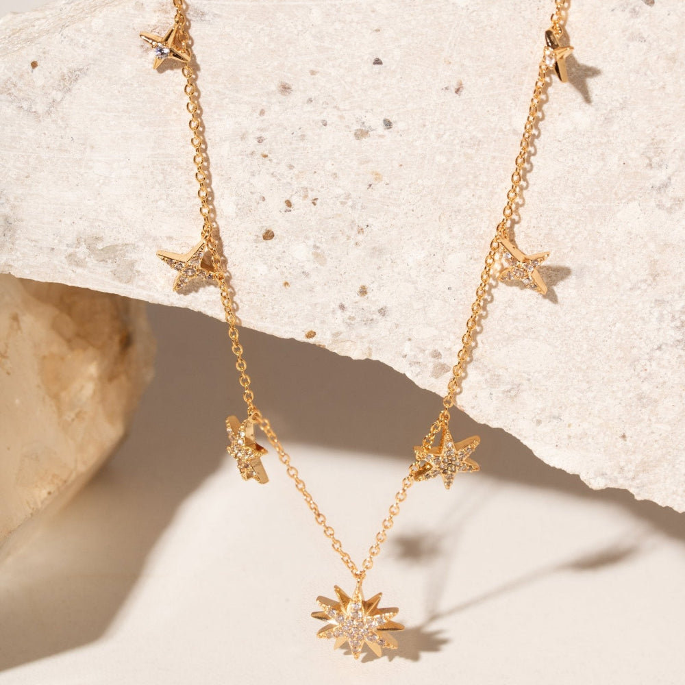 A gold-toned necklace with multiple starburst and bird charms, displayed draped over a textured light beige stone slab.