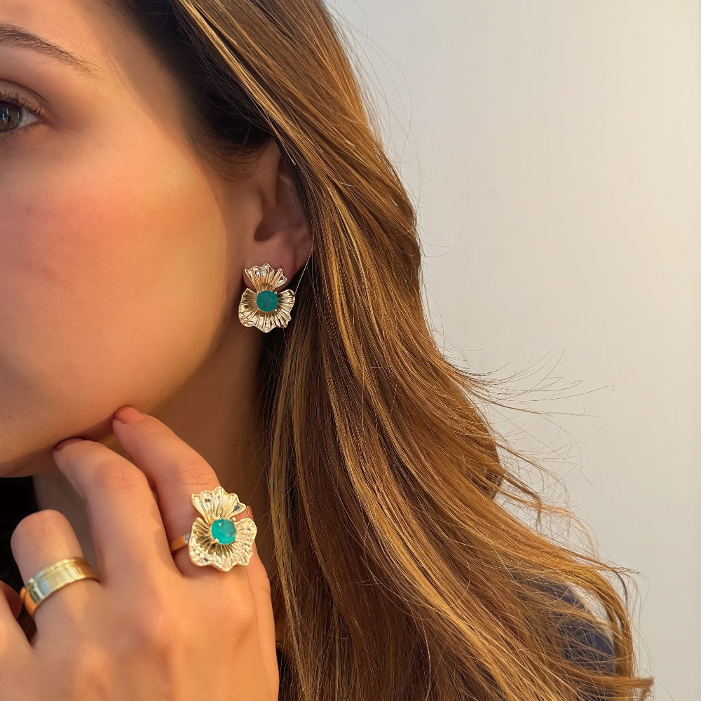 A close-up lifestyle shot of a woman wearing matching gold flower-shaped stud earrings and a ring, each featuring a central turquoise cabochon stone.