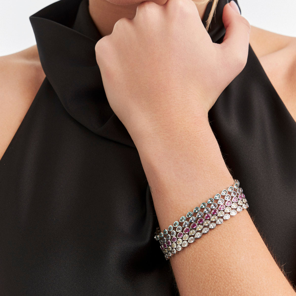 A close-up of a model's wrist wearing a silver-toned multi-band bracelet adorned with clear round stones and rows of small pink gemstones, paired with a black satin top.