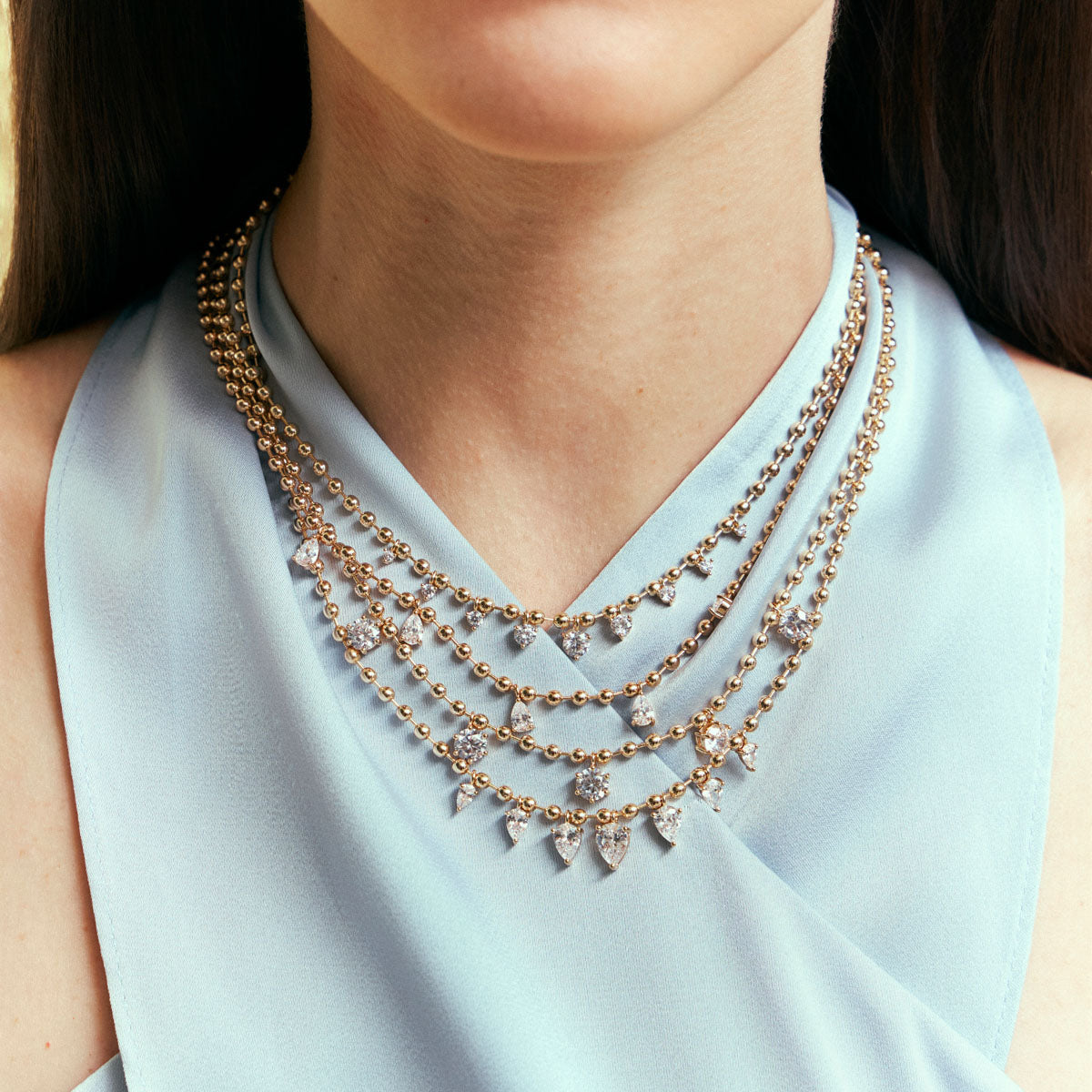 A close-up of a model wearing a multi-strand rose gold statement necklace with clear crystal pendants, styled with a light blue satin top.