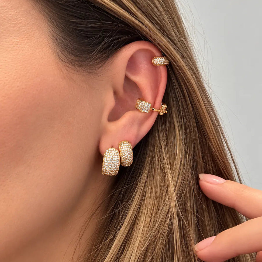 A close-up of a model's ear wearing multiple gold-toned ear cuff and hoop earrings covered in small clear stones, with her hair and hand visible against a light gray background.