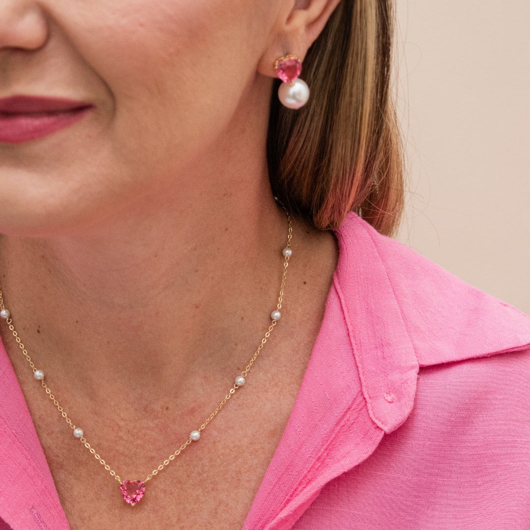 A close-up lifestyle shot of a woman wearing a gold necklace with a pink heart-shaped crystal pendant and matching drop earrings with pink crystals and white pearls, paired with a bright pink shirt against a solid beige background.
