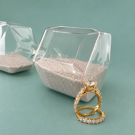 Two gold rings with clear gemstones, one standing upright and the other lying flat, displayed on a teal background with clear geometric glass containers containing light gray beads.