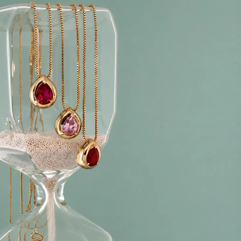 Three gold teardrop pendant necklaces with red, pink, and red gemstones are displayed on a clear glass hourglass filled with sand against a solid teal background.