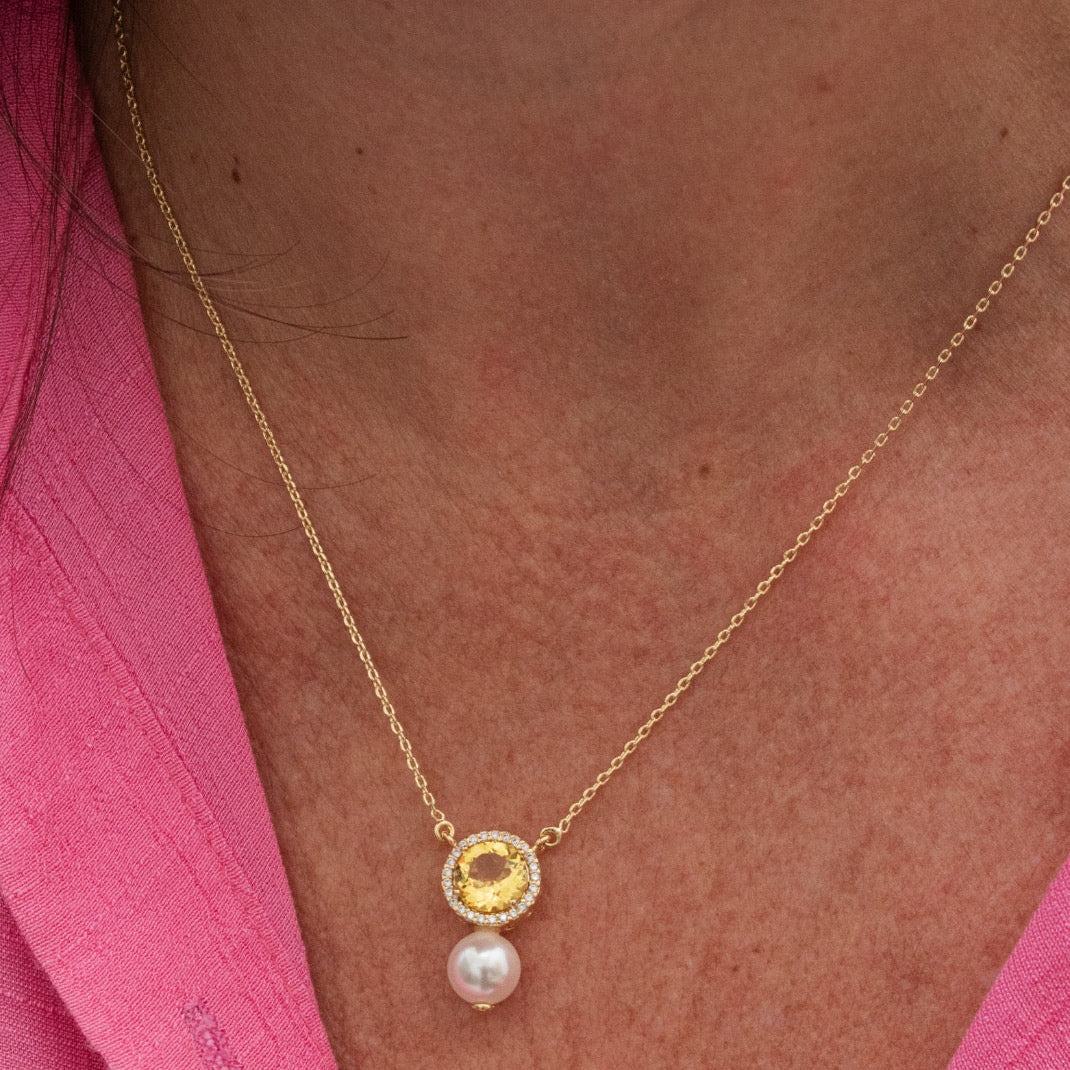 A close-up of a person wearing a delicate gold chain necklace with a circular gold pendant and a dangling white pearl, styled with a pink top.