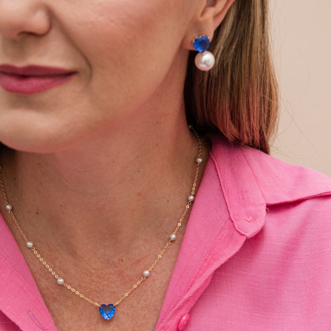 A close-up lifestyle shot of a woman wearing a gold necklace with a blue heart-shaped crystal pendant and matching drop earrings with blue and white stones, paired with a pink collared shirt.