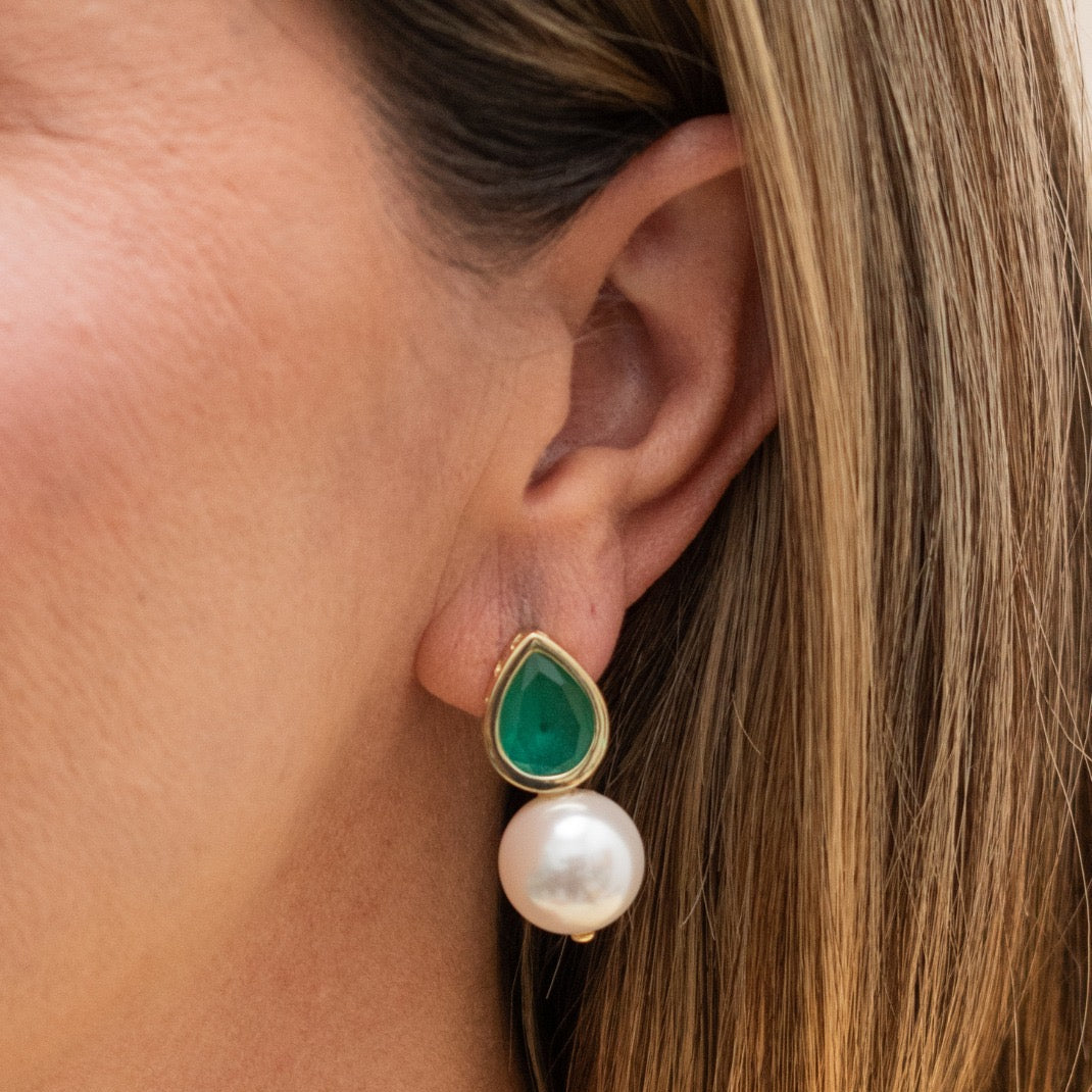 A close-up of a woman's ear wearing a gold drop earring with a green teardrop-shaped stone and a white pearl drop.