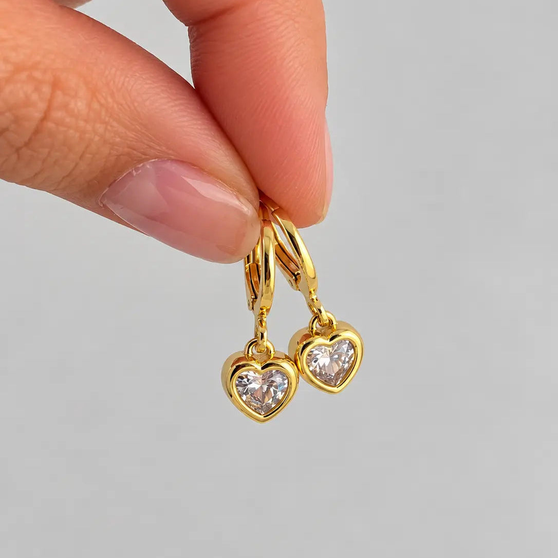 A close-up of a hand holding a pair of gold heart-shaped hoop earrings with clear heart-shaped gemstones against a light gray background.