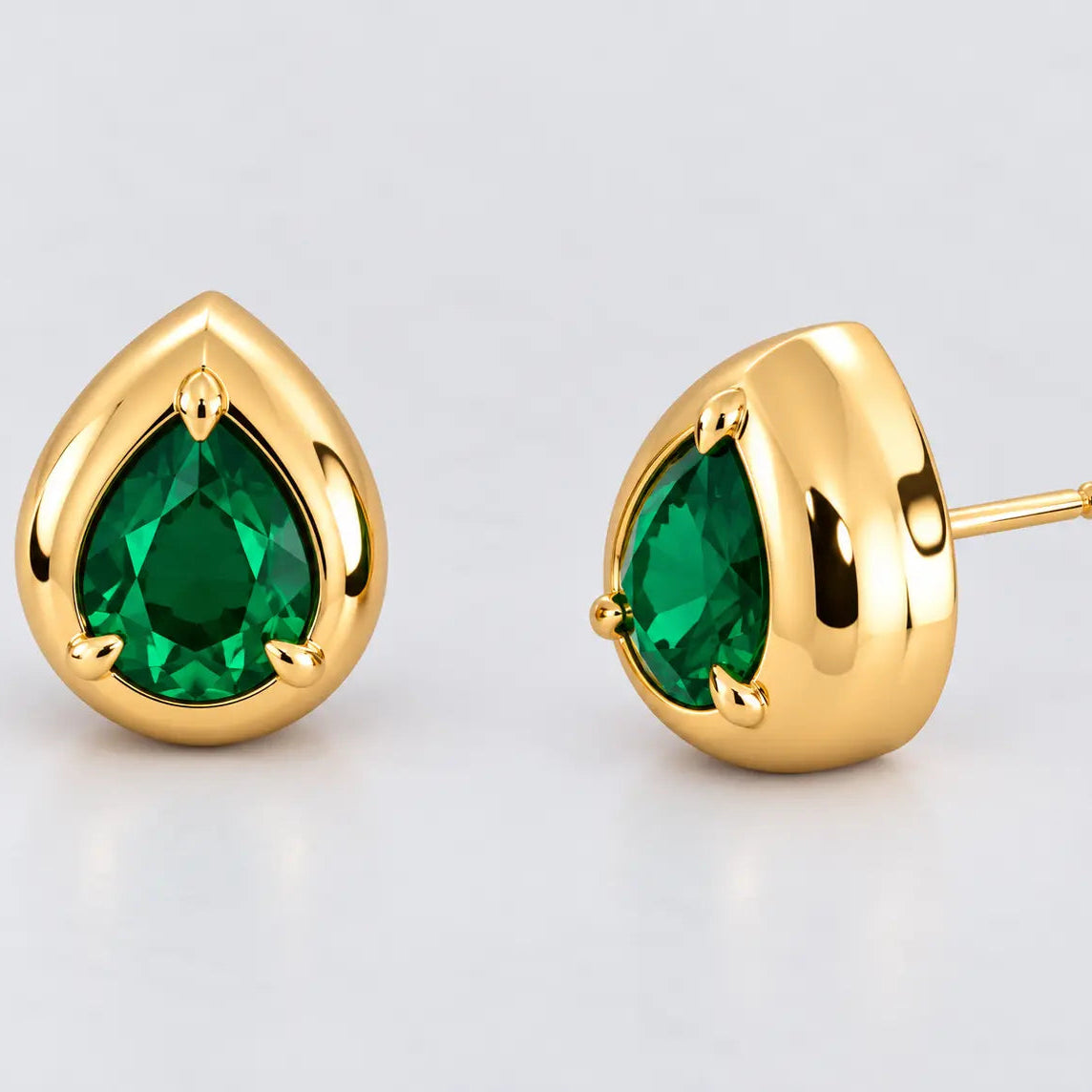 A pair of gold teardrop stud earrings with green teardrop-cut gemstones, shown on a light gray reflective surface. One earring is shown from the front and the other from the side profile, revealing the post.